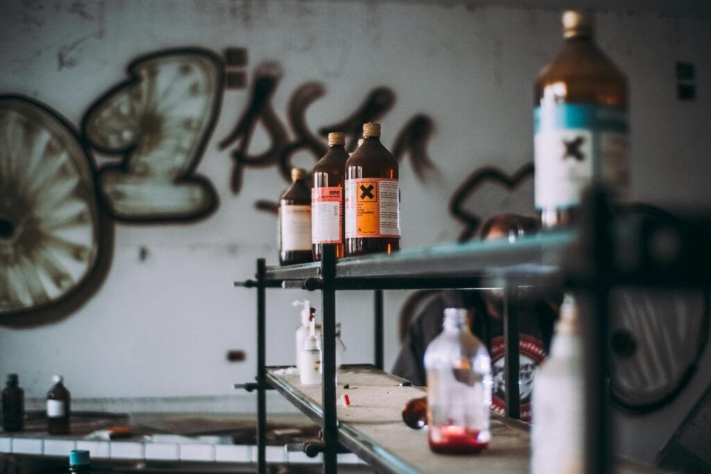 Displays several bottles on shelves in an abandoned setting, some marked with a hazard symbol, signifying toxic substances.