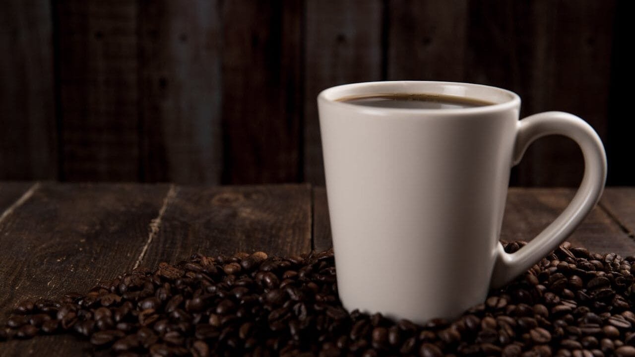 A white mug filled with coffee sitting on a wooden table surrounded by coffee beans.