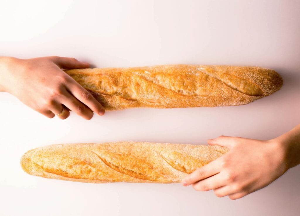 Two hands reaching for two baguettes, which are long, thin loaves of French bread.