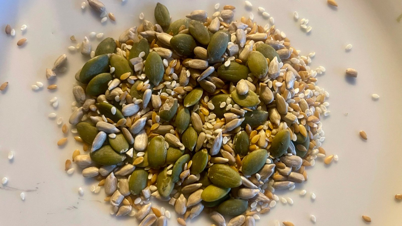 A mix of seeds, likely an "Organic Omega Four Seed Mix" which typically contains pumpkin seeds, sunflower seeds, golden linseeds (flax seeds), and sesame seeds.